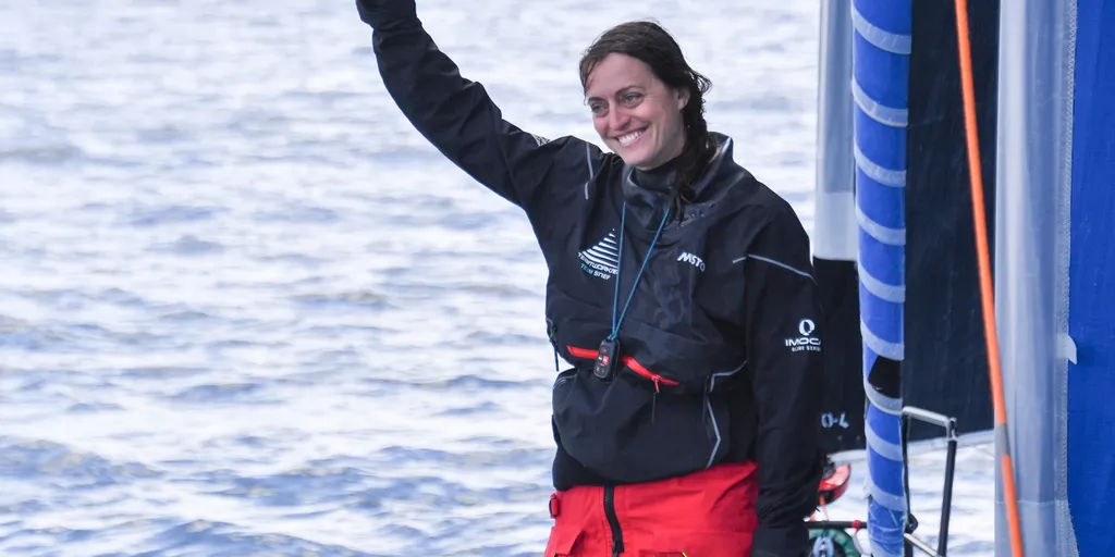 Justine Mettraux, souriante, en veste de quart noire et pantalon de ciré rouge, lève le pouce sur le pont de son IMOCA, la mer en arrière-plan.
