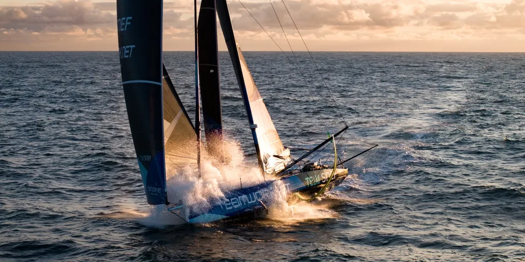 Aerial view of the IMOCA TeamWork - Team SNEF, with its blue hull and dark sails, speeding offshore in a burst of spray under an evening sky.