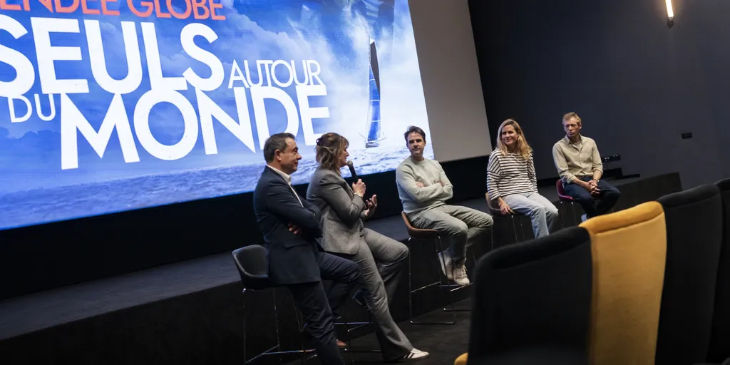 Five speakers seated on high stools on the stage of a cinema, in front of a screen displaying “Vendée Globe – Seuls autour du monde”. A woman in a grey suit speaks into a microphone, flanked by two men and, on the right, a female skipper in a striped top and a male skipper in a beige shirt.