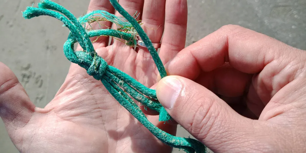 Main tenant un cordage de pêche vert, noué et effiloché, couvert de sable, trouvé sur une plage.