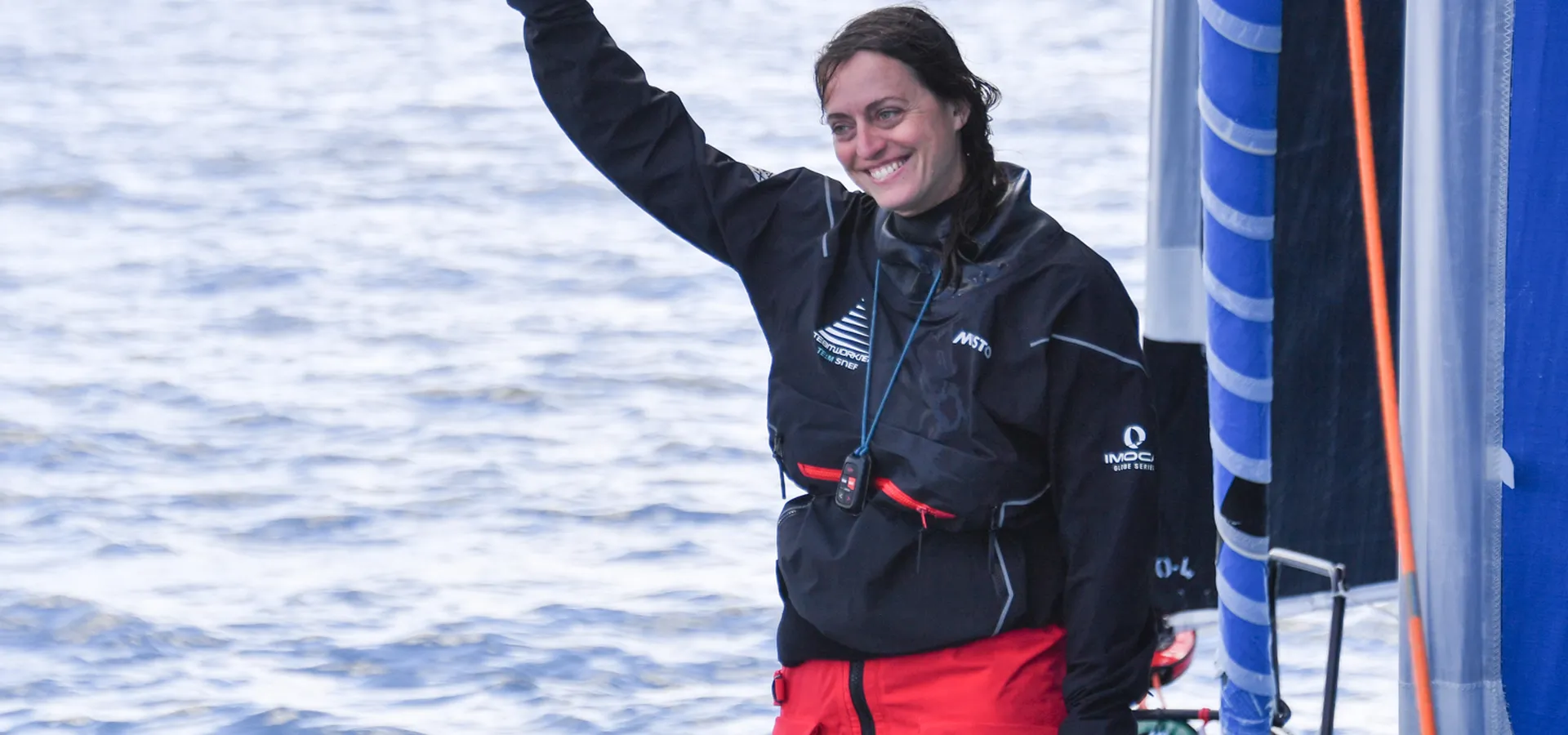 Justine Mettraux, souriante, en veste de quart noire et pantalon de ciré rouge, lève le pouce sur le pont de son IMOCA, la mer en arrière-plan.