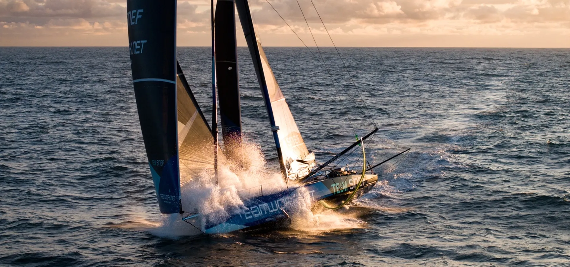 Vue aérienne de l’IMOCA TeamWork - Team SNEF, coque bleue et voiles sombres, filant au large dans une gerbe d’écume sous un ciel de fin de journée.