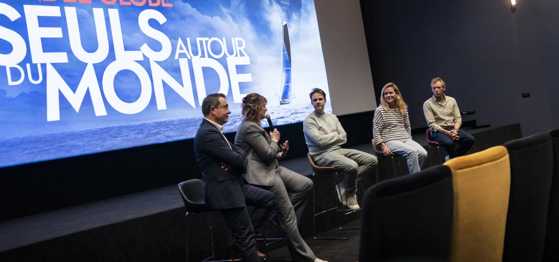 Cinq intervenants assis sur des tabourets hauts sur la scène d’une salle de cinéma, devant un écran affichant « Vendée Globe – Seuls autour du monde ». Une femme en tailleur gris parle au micro, entourée de deux hommes et, à droite, d’une skippeuse en marinière et d’un skipper en chemise beige.