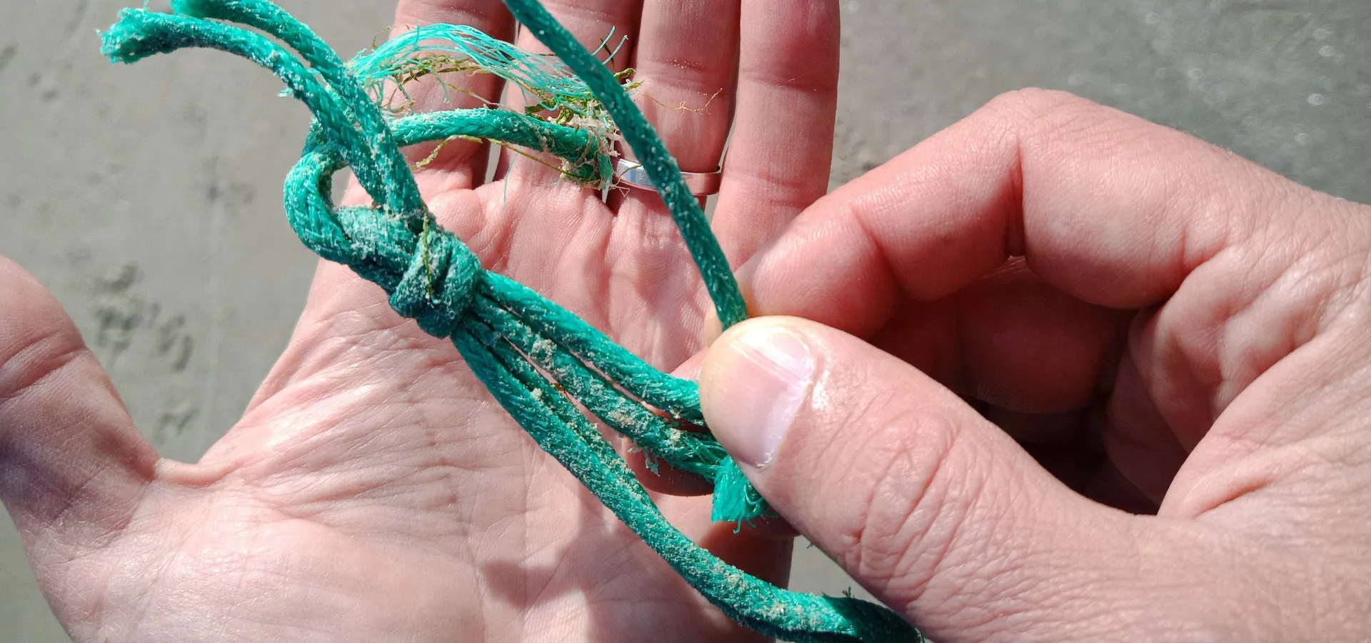Main tenant un cordage de pêche vert, noué et effiloché, couvert de sable, trouvé sur une plage.