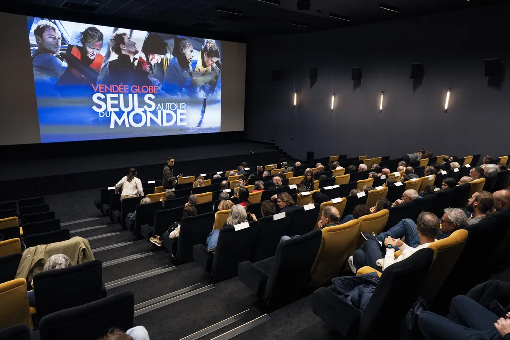 High-angle view of a cinema with black and mustard-yellow seats as the audience takes their places. On the big screen, the poster for the series “Vendée Globe – Seuls autour du monde” shows the faces of several skippers at sea.