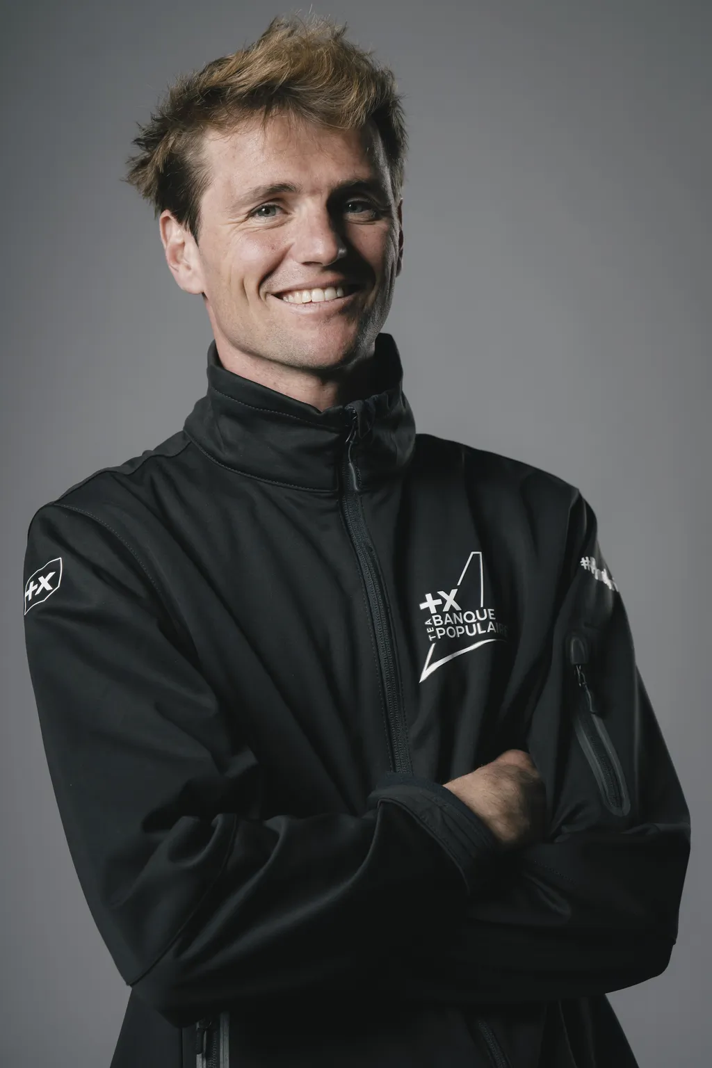 Portrait en studio de Loïs Berrehar, souriant, bras croisés, vêtu d’une veste softshell noire floquée du logo Team Banque Populaire, sur fond gris uni.