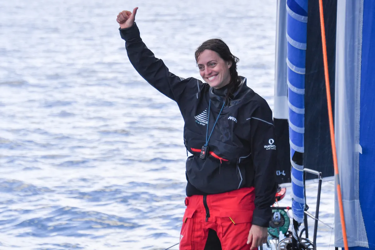 A smiling Justine Mettraux, wearing a black sailing jacket and red foul-weather trousers, gives a thumbs-up on the deck of her IMOCA, with the sea in the background.