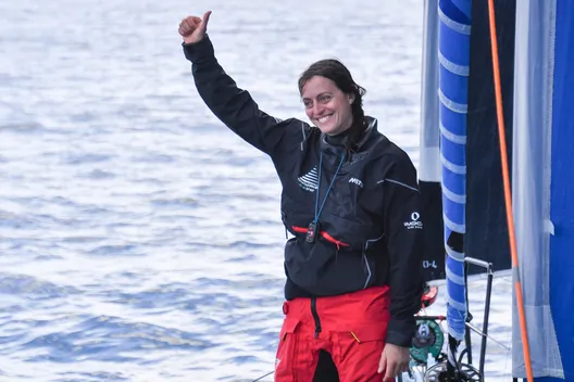 A smiling Justine Mettraux, wearing a black sailing jacket and red foul-weather trousers, gives a thumbs-up on the deck of her IMOCA, with the sea in the background.