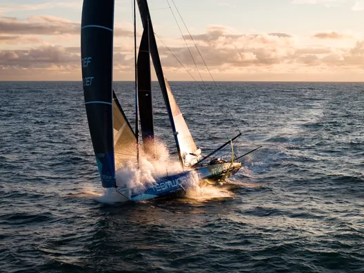 Aerial view of the IMOCA TeamWork - Team SNEF, with its blue hull and dark sails, speeding offshore in a burst of spray under an evening sky.