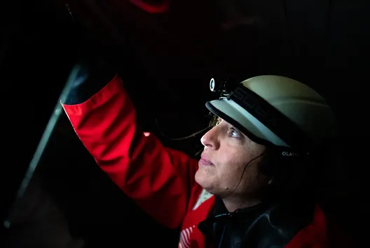Justine Mettraux, wearing a white helmet with a head torch and a red foul-weather jacket, raises her arm and looks upward in the dim light of her boat.