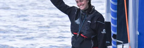 Justine Mettraux, souriante, en veste de quart noire et pantalon de ciré rouge, lève le pouce sur le pont de son IMOCA, la mer en arrière-plan.