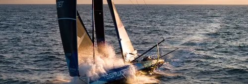 Aerial view of the IMOCA TeamWork - Team SNEF, with its blue hull and dark sails, speeding offshore in a burst of spray under an evening sky.