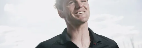 Loïs Berrehar, souriant, en polo noir Team Banque Populaire, photographié à contre-jour, le regard levé vers un ciel nuageux, avec des mâts de voiliers en arrière-plan.