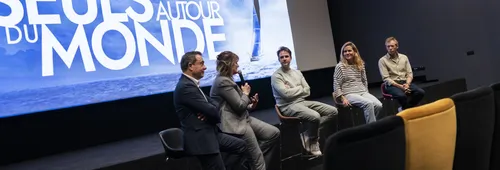 Five speakers seated on high stools on the stage of a cinema, in front of a screen displaying “Vendée Globe – Seuls autour du monde”. A woman in a grey suit speaks into a microphone, flanked by two men and, on the right, a female skipper in a striped top and a male skipper in a beige shirt.